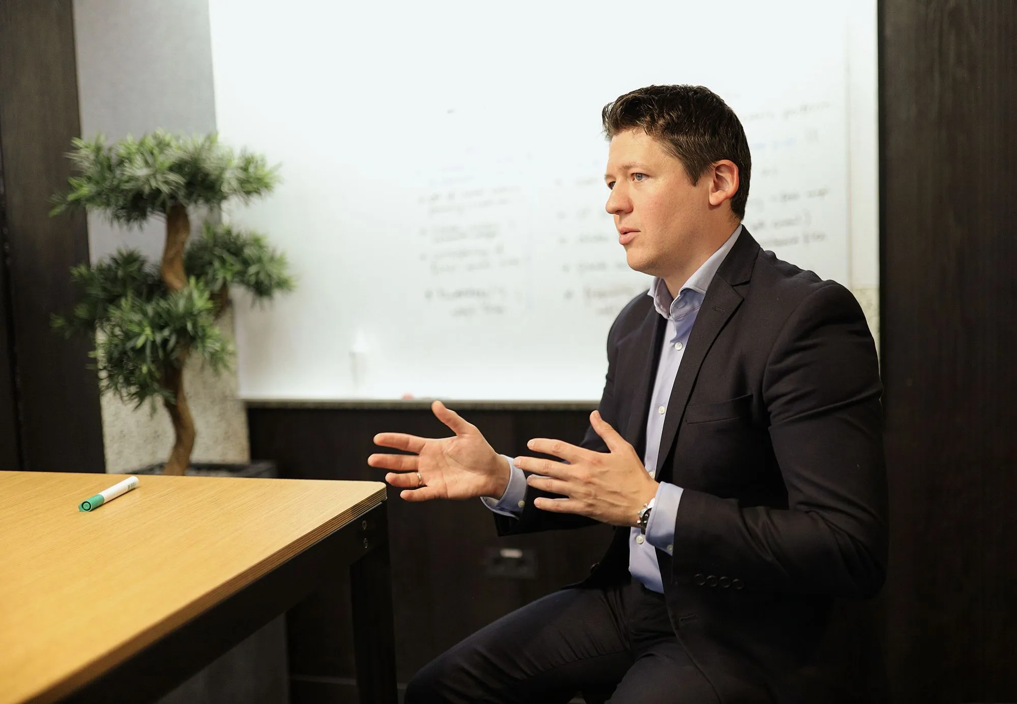 Alex Kukharenko at a table, explaining something with both hands, a whiteboard behind him.